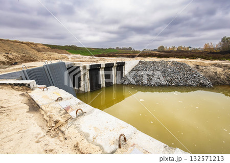 A construction area shows a water management system with gates and a small pond surrounded by gravel and earth under a cloudy sky 132571213