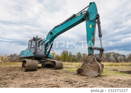 A large excavator operates in an open field, digging into the dirt on a cloudy day. Surrounding trees hint at an ongoing construction project 132571227