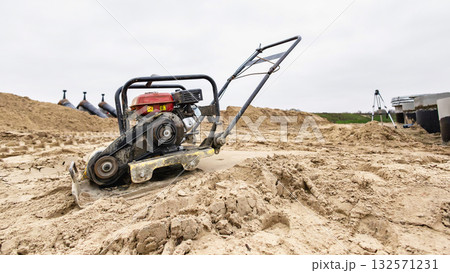 A compacting machine uses vibrations to compress sand on a construction site. Workers prepare the ground for building amidst cloudy skies in a rural area 132571231