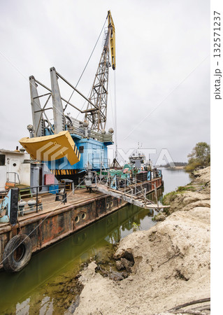 A large crane is positioned on a barge at a river construction site, with equipment lined up for operation on a cloudy day 132571237