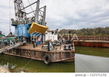 A large barge with construction equipment is docked at a waterway, surrounded by trees, under cloudy skies. Workers can be seen preparing for tasks A large barge with construction equipment is docked at a waterway, surrounded by trees, under cloudy skies. Workers can be seen preparing for tasks 132571238
