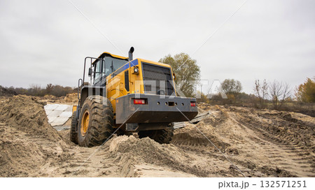 A large yellow tractor is digging in sandy soil at a construction site. The cloudy sky and autumn trees create a somber backdrop for the activity A large yellow tractor is digging in sandy soil at a construction site. The cloudy sky and autumn trees create a somber backdrop for the activity 132571251