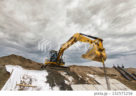 A yellow excavator lifts soil at a construction site surrounded by piles of dirt, with dark clouds overhead during the late afternoon A yellow excavator lifts soil at a construction site surrounded by piles of dirt, with dark clouds overhead during the late afternoon 132571256