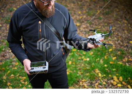 A person stands in a lush green area, holding a drone while gripping a controller. The sunlight filters through trees, creating a serene atmosphere for exploration. 132572730