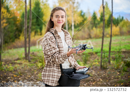 In a beautiful forest during autumn, a smiling young woman stands proudly holding a drone. Colorful trees surround her, showcasing the season's vibrant hues. 132572732