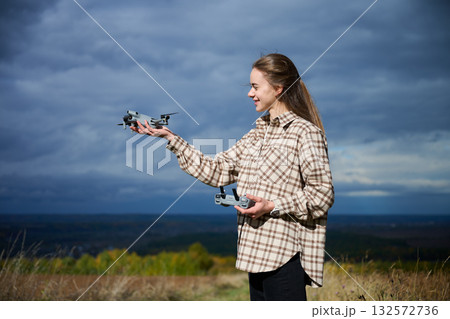 A young woman stands in a grassy area, holding drones in both hands as she prepares to fly them. The clouds create a moody backdrop, enhancing the adventurous scene. 132572736