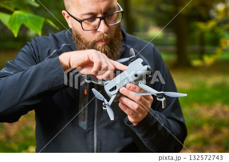 In a serene park filled with falling leaves, a man with glasses carefully adjusts his drone, preparing for a flight. The vibrant nature surrounds him. 132572743