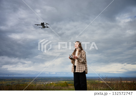 A young woman stands on a hilltop with a remote control, flying her drone above an expansive vista. The clouds create a dramatic backdrop as she smiles joyfully. A young woman stands on a hilltop with a remote control, flying her drone above an expansive vista. The clouds create a dramatic backdrop as she smiles joyfully. 132572747