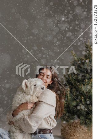 Young woman embraces fluffy Westie near evergreen as snow falls in studio, cozy winter holiday lifestyle portrait Young woman embraces fluffy Westie near evergreen as snow falls in studio, cozy winter holiday lifestyle portrait 132573278