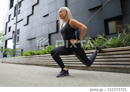 Woman performing a lunge stretch outdoors near modern architecture building 132573725
