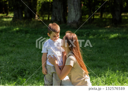 Young Caucasian mother laughs giggling son across sunny park lawn. Dynamic image radiates pure joy, energy, and fun of outdoor playtime together. 132576589