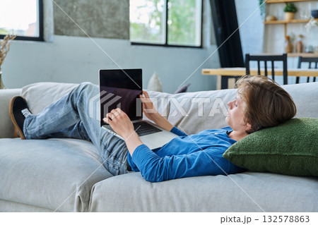 Serious teenage boy lying at home on the couch using laptop Serious teenage boy lying at home on the couch using laptop 132578863