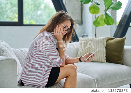 Sad serious middle aged woman with smartphone sitting on sofa 132578878