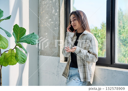 Serious teenage female standing at home talking on smartphone Serious teenage female standing at home talking on smartphone 132578909