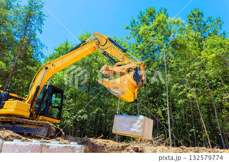 Excavator operates in laying large stone block during construction retaining wall landscaping work Excavator operates in laying large stone block during construction retaining wall landscaping work 132579774