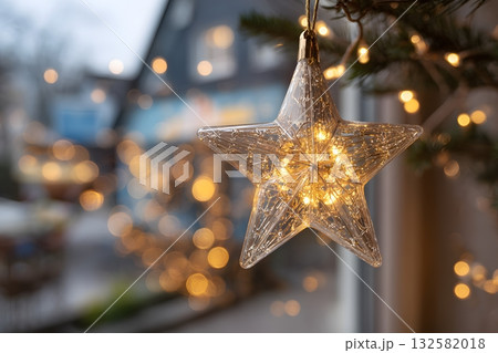 A clear Christmas star ornament glows with warm lights, hanging near a blurry outdoor scene. 132582018