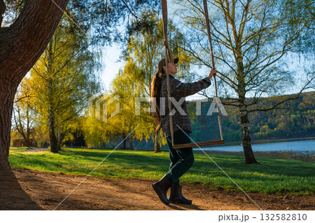Woman standing by wooden swing under green spring trees near lake on sunny day in Czech Republic 132582810