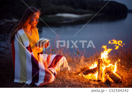 Young blond girl sitting near campfire on river bank and dreaming of something. Young girl drinking warm tea from her cup, admiring the calm evening near the river 132584242