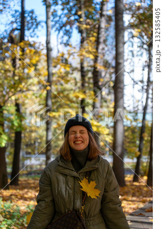 Woman in a green jacket and black beanie smiles joyfully in an autumn park with yellow leaves and sunlight filtering through the trees Woman in a green jacket and black beanie smiles joyfully in an autumn park with yellow leaves and sunlight filtering through the trees 132585405