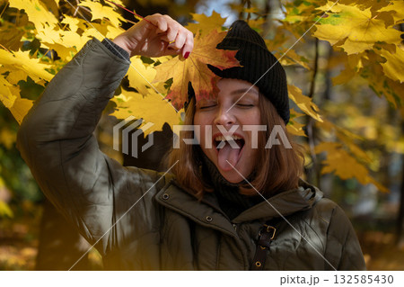 Playful woman in a green jacket and black beanie holds a yellow maple leaf to her face and sticks out her tongue in an autumn park 132585430