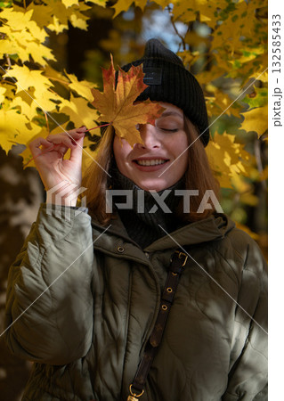 Woman in a green jacket and black beanie smiles with eyes closed, holding a yellow maple leaf over her face in an autumn park 132585433