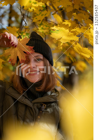 Smiling woman in a green jacket and black beanie holds a yellow maple leaf near her face in a golden autumn forest Smiling woman in a green jacket and black beanie holds a yellow maple leaf near her face in a golden autumn forest 132585437