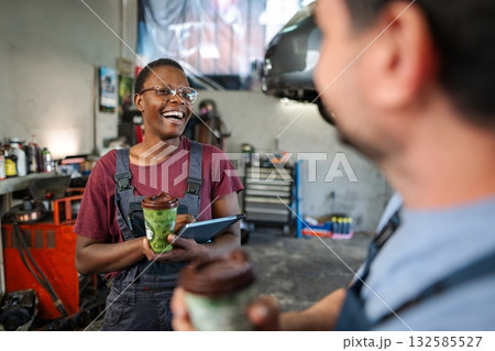 Cheerful african american female mechanic laughing with colleague during coffee break at auto repair shop Cheerful african american female mechanic laughing with colleague during coffee break at auto repair shop 132585527