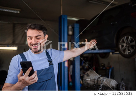 Happy mechanic showing a lifted car while using a smartphone in 132585532
