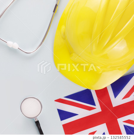 Medical care and insurance of workers in Great Britain. Flag of Great Britain, stethoscope and yellow hardhat on a blue background. Labor safety and right at Workplace in the UK Medical care and insurance of workers in Great Britain. Flag of Great Britain, stethoscope and yellow hardhat on a blue background. Labor safety and right at Workplace in the UK 132585552