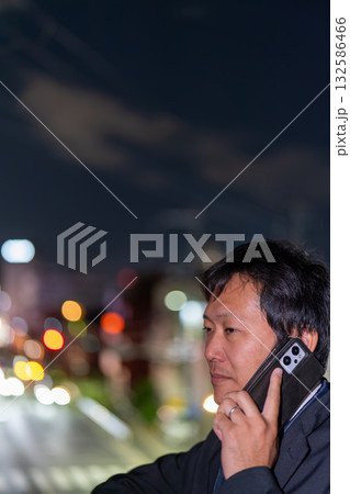 街の夜景を背に緊急対応するサラリーマン、残業帰りに取引先へ連絡する男性社員 街の夜景を背に緊急対応するサラリーマン、残業帰りに取引先へ連絡する男性社員 132586466