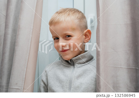 Boy smiles shyly while peeking between gray curtains warm indoor moment full of curiosity 132586945