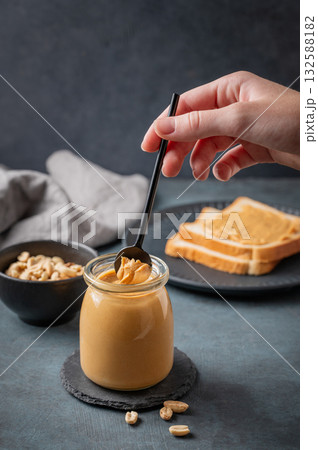 A hand holds a spoon with creamy peanut butter in a jar on a dark background with toast bread A hand holds a spoon with creamy peanut butter in a jar on a dark background with toast bread 132588182