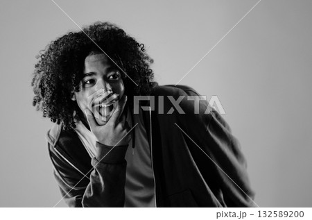 Young man with curly hair is making a funny face and sticking out his tongue while holding his chin 132589200