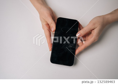 Top view of woman removing broken tempered glass screen protector from smartphone, revealing cracked protection film on white table. Concept of mobile phone maintenance and service. 132589463