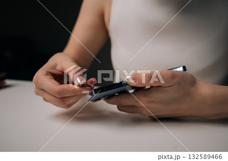 Close-up view of woman removing damaged tempered glass screen protector from smartphone with fingers, sitting at white table. Concept of mobile phone maintenance and service. 132589466