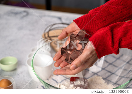 Close-up of hands holding a gingerbread man cookie cutter above a bowl of flour. Holiday baking preparation. Christmas cookie recipe, festive kitchen concept, home baking 132590008
