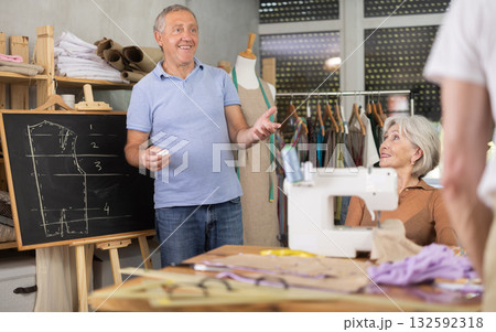 Sewing school senior man teacher conducts lesson for elderly students, draws pattern on black-board Sewing school senior man teacher conducts lesson for elderly students, draws pattern on black-board 132592318