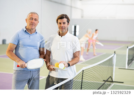 Portrait of two men pickleball players with rackets and ball standing on an indoor court Portrait of two men pickleball players with rackets and ball standing on an indoor court 132592329