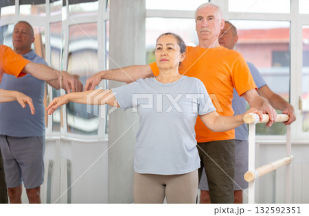 Mature woman in sportswear participating 2nd position of ballet stance at barre with group in choreography studio Mature woman in sportswear participating 2nd position of ballet stance at barre with group in choreography studio 132592351