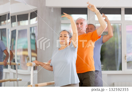 Happy smiling old woman dressed in sportswear enjoying ballet choreography, standing in fourth position at barre during group class in dance salon 132592355