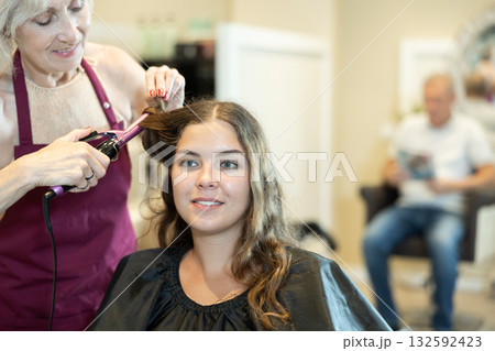 Female hairdresser makes curls the hair of young woman using curling iron 132592423