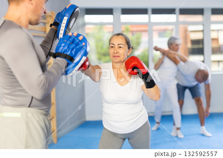 Senior lady and male couch are boxing in gym, man helps student to work out force of blow. Senior lady and male couch are boxing in gym, man helps student to work out force of blow. 132592557