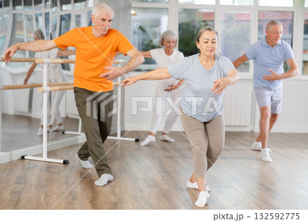 Experienced mature female ballet dancer, dressed in sportswear executing dance moves with focused precision among group of aged people rehearsing in ballet studio Experienced mature female ballet dancer, dressed in sportswear executing dance moves with focused precision among group of aged people rehearsing in ballet studio 132592775