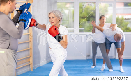 Old woman training boxing kicks on punch mitts held by instructor 132592837