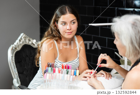 Friendly elderly woman working in beauty salon as manicure master shaping the nails of young female client with nail file Friendly elderly woman working in beauty salon as manicure master shaping the nails of young female client with nail file 132592844