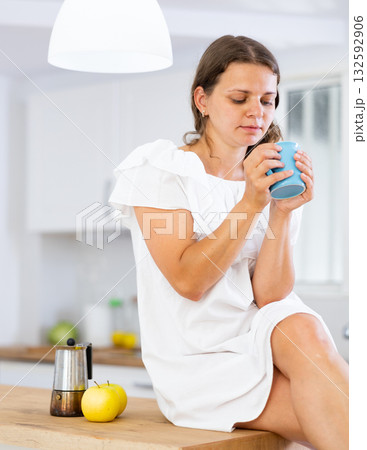 Woman sitting on countertop at kitchen and looking in cup Woman sitting on countertop at kitchen and looking in cup 132592906