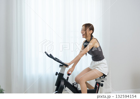 Young woman enjoys happy home activity, cycling on stationary bike for fitness, feeling energetic and enthusiastic during healthy workout with headphones, promoting good wellness lifestyle Young woman enjoys happy home activity, cycling on stationary bike for fitness, feeling energetic and enthusiastic during healthy workout with headphones, promoting good wellness lifestyle 132594154