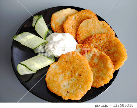 Potato pancakes served with sour cream and cucumber slices on a black plate. 132599319
