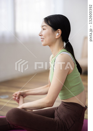ヨガマットの上でストレッチをするワイヤレスイヤフォンをつけたミドル女性 132600392