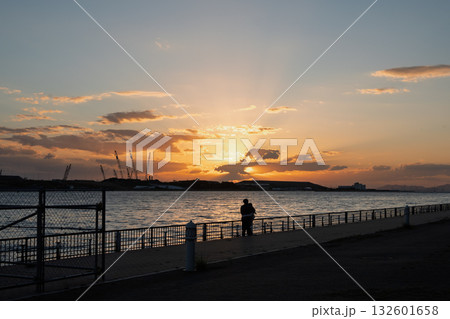 大阪湾の夕暮れと対岸の万博会場工事現場を見つめるカップルのシルエット(デート・未来・黄昏) 大阪湾の夕暮れと対岸の万博会場工事現場を見つめるカップルのシルエット(デート・未来・黄昏) 132601658